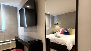 Una habitación de hotel moderna con un espejo grande, un televisor de pantalla plana en la pared, una cama con almohadones de colores, un otoman rojo y suelo de madera.