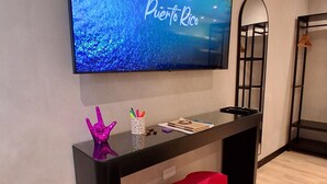 Una habitación moderna con un televisor de pantalla plana grande que muestra 'Puerto Rico', un escritorio de madera oscura con un reposapiés rojo y un espejo con marco negro.