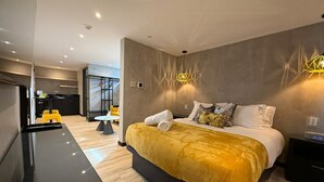 Une chambre d’hôtel moderne avec un grand lit, un petit bureau et un coin cuisine à l’arrière-plan.