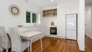 A compact kitchen with white cabinetry, a built-in oven, a refrigerator, and a dining area with wicker chairs. - 2br - Little Opulence Beach Villa on Marine (Kingscliff)