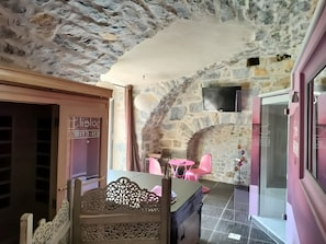 Une pièce avec un mur en pierre, un lit avec une tête de lit, une chaise rose, une table et un téléviseur fixé au mur.