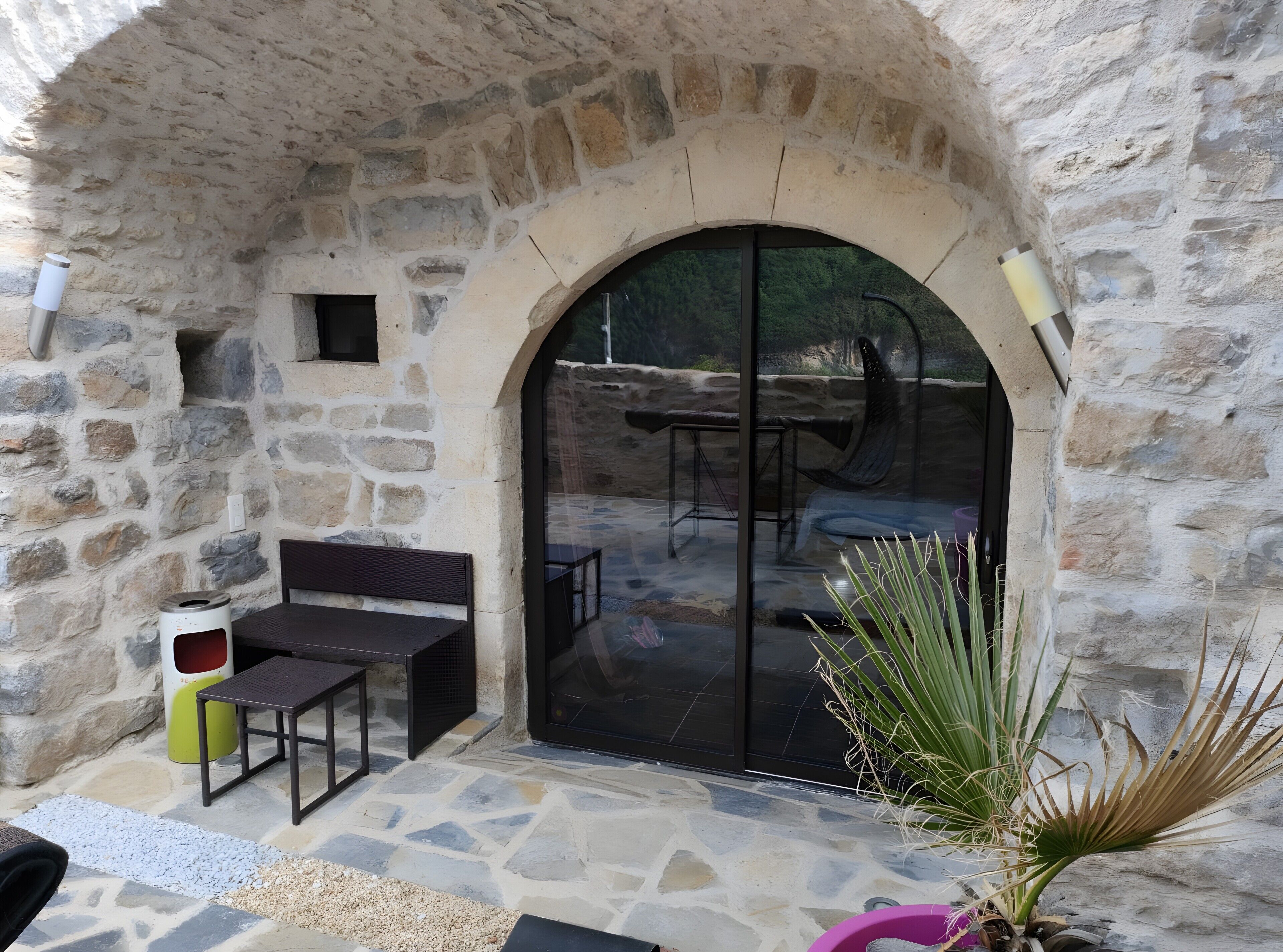 Une terrasse extérieure avec un sol en pierre, un espace salon comprenant un banc et une table, et une grande porte vitrée donnant sur un espace intérieur.