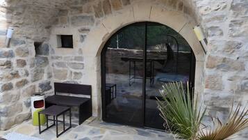 Une terrasse extérieure avec un sol en pierre, un espace salon comprenant un banc et une table, et une grande porte vitrée donnant sur un espace intérieur.