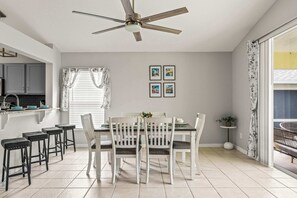A dining area with a white table, four chairs, and a ceiling fan. There are framed pictures on the wall and a window with curtains. - The Sunny Spot Home for 8 (Davenport)