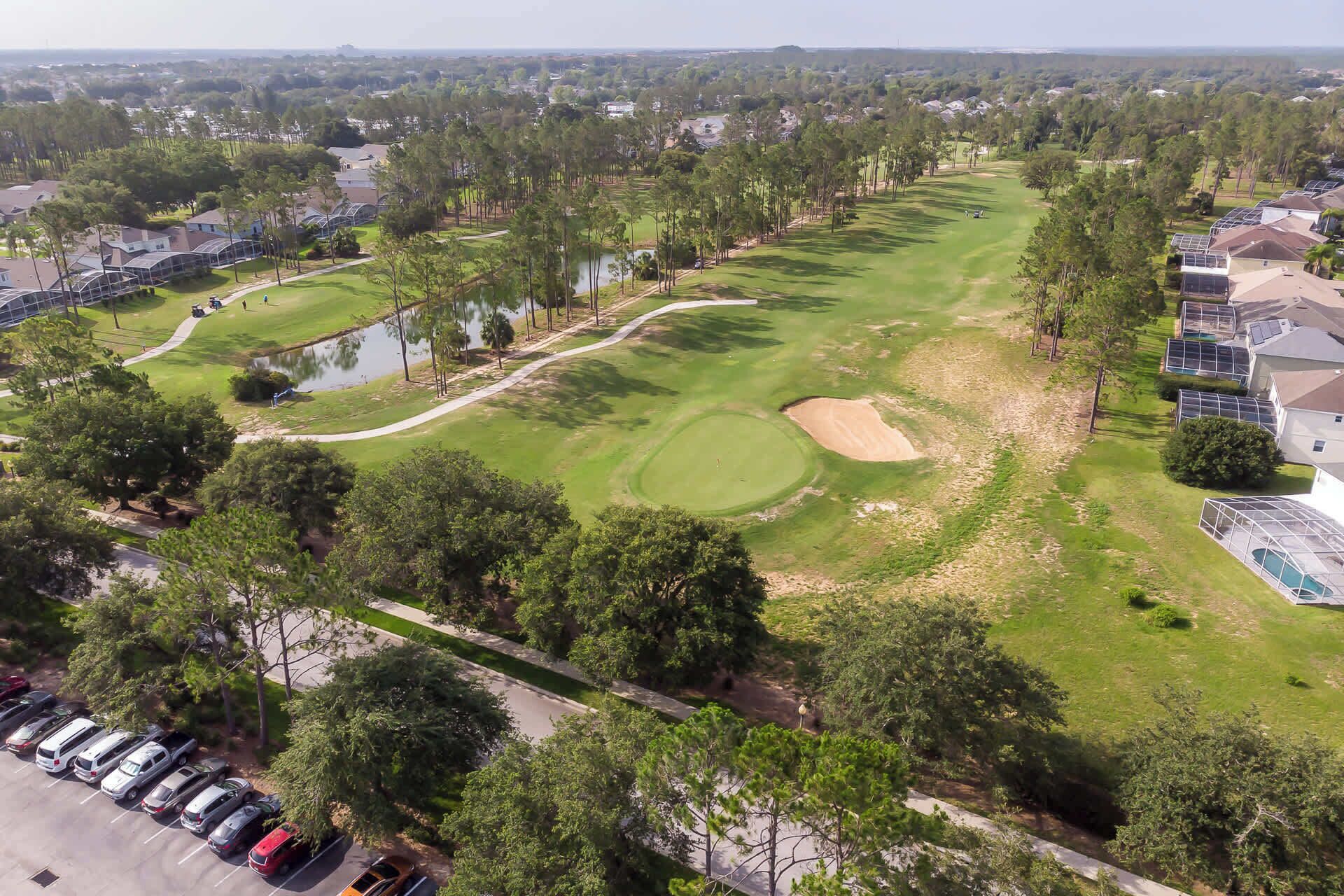 An aerial view of a golf course with a pond, surrounded by residential housing and a parking area.