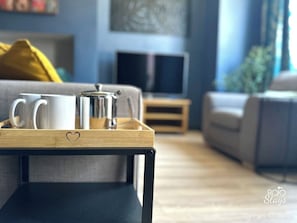 A modern living room with a sofa, a coffee table with a tray holding a coffee pot and cups, and a television on a stand.