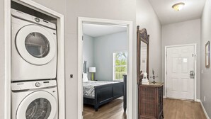 Un cuarto de lavado con lavadora y secadora Samsung, un dormitorio con cama y mesita de noche, y un pasillo con una puerta y un espejo.