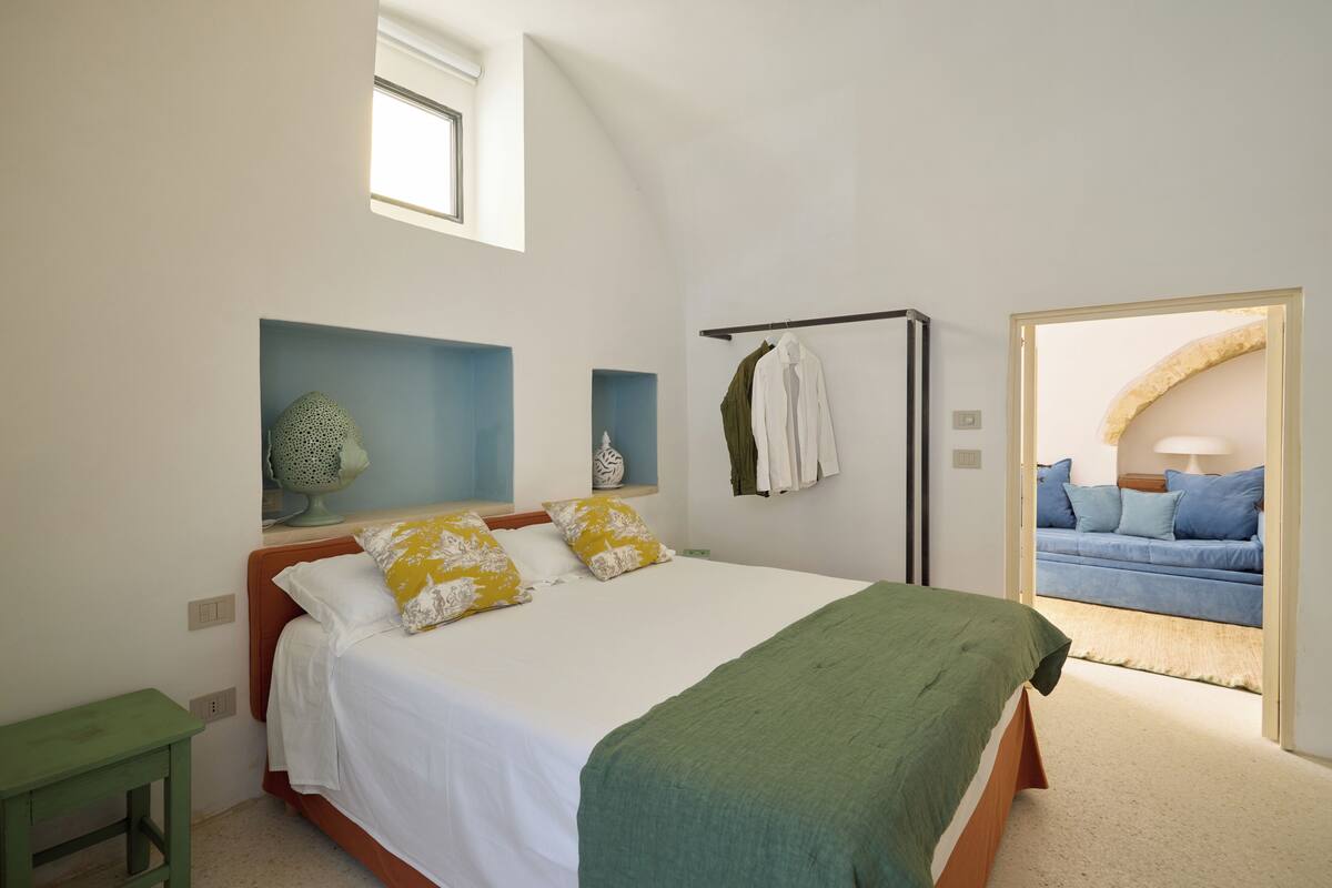 A bedroom with a bed, a green side table, a wall-mounted drying rack, and a view into another room with a sofa.