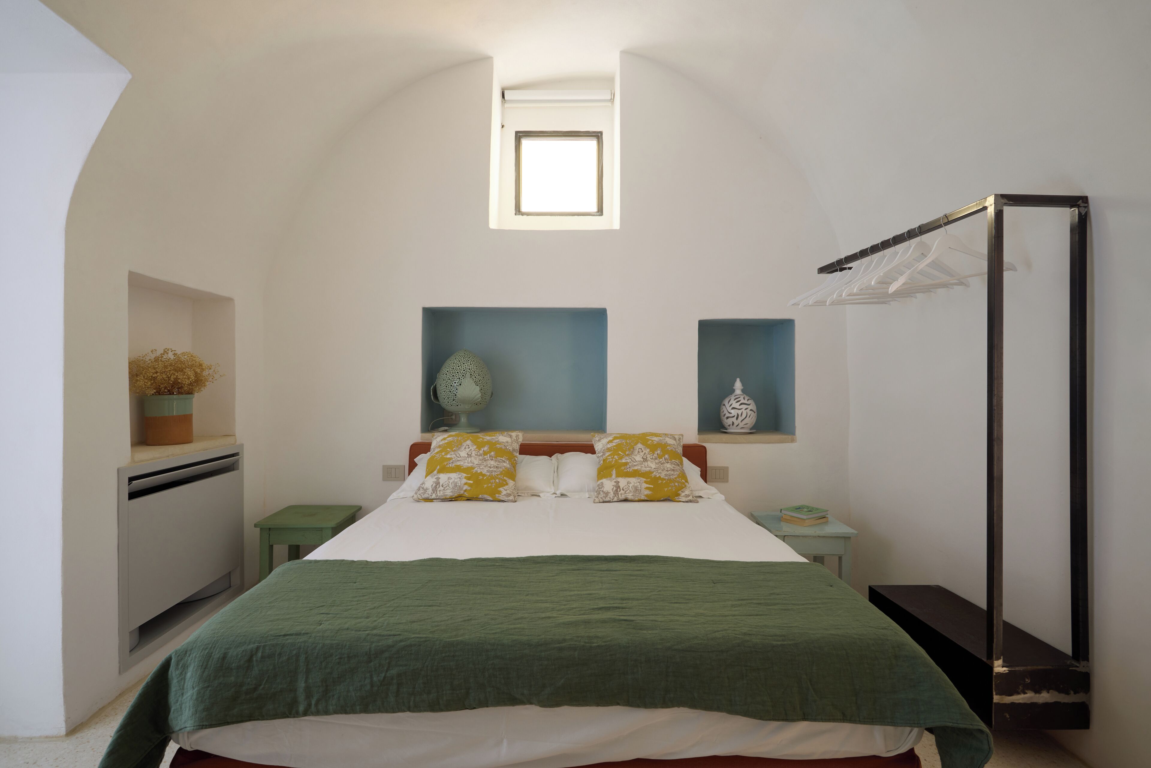 A bedroom with a bed, a small table, a nightstand, a wardrobe, a window, and a shelf with decorative items.