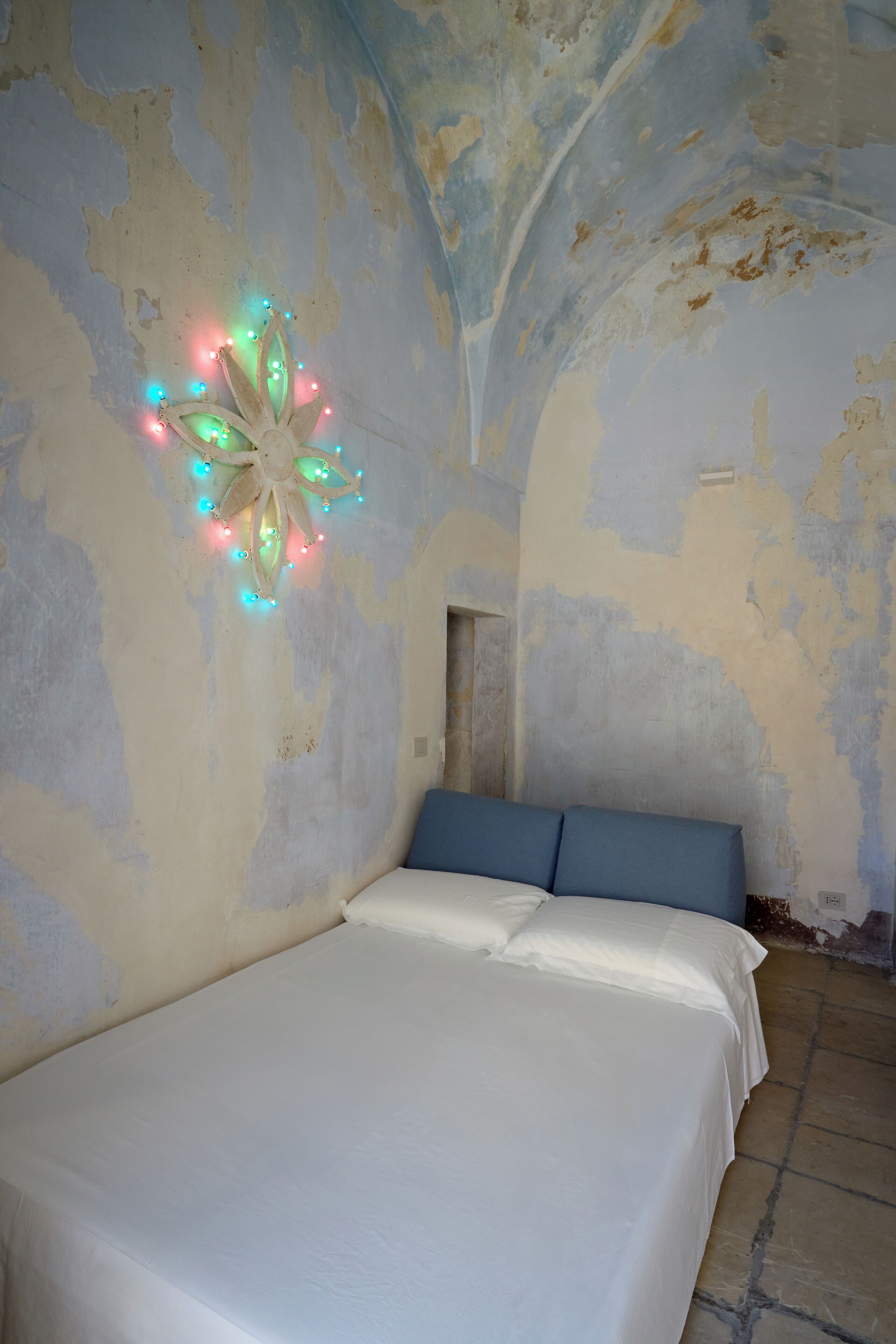 Un dormitorio con cama, una lámpara decorativa y pintura descascarada en la pared.