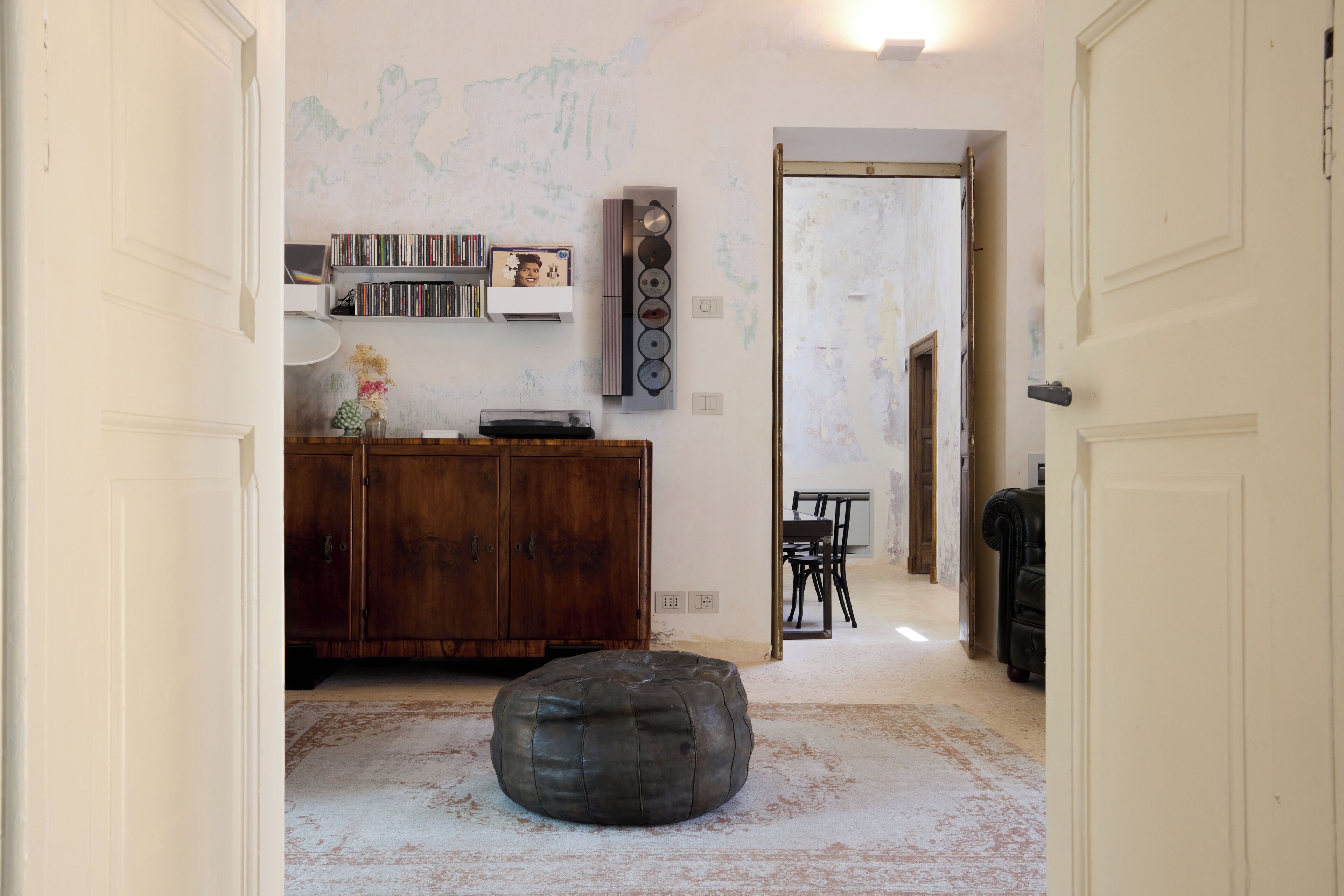 Un pasillo con un armario de madera, estanterías, un otoman redondo y un comedor con sillas.