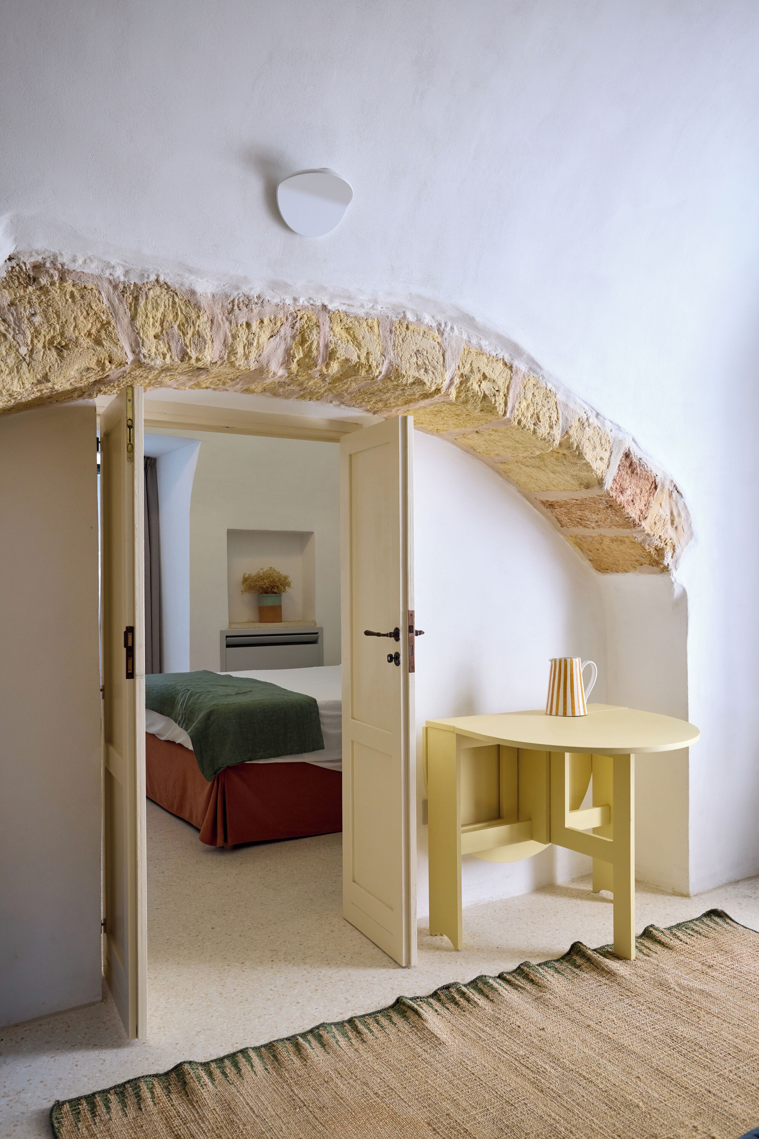 A room with a high, textured ceiling, a bed with a green blanket, a yellow table with a striped mug, and a small plant on a shelf.