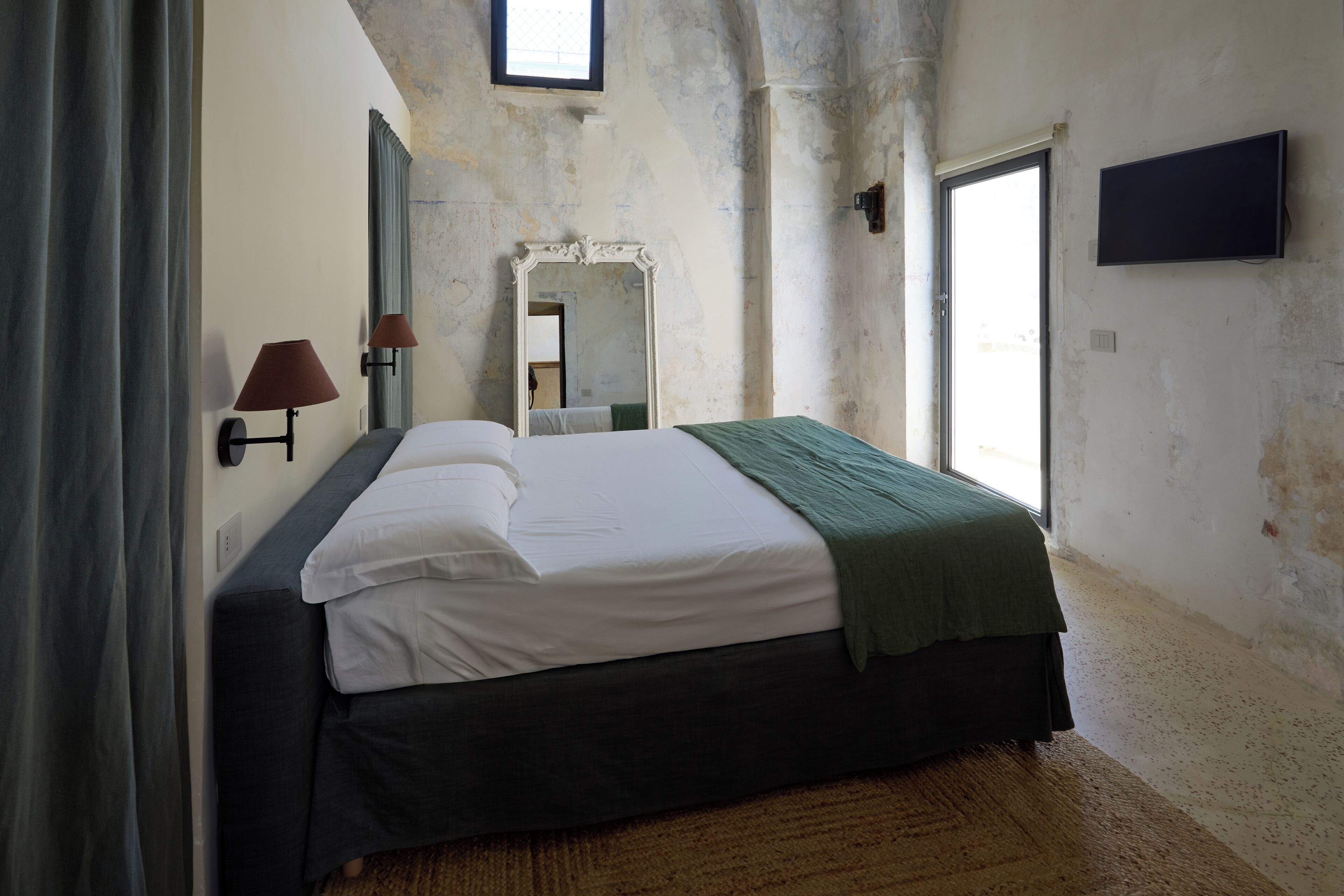 A bedroom with a bed, a mirror, a TV, and a window.