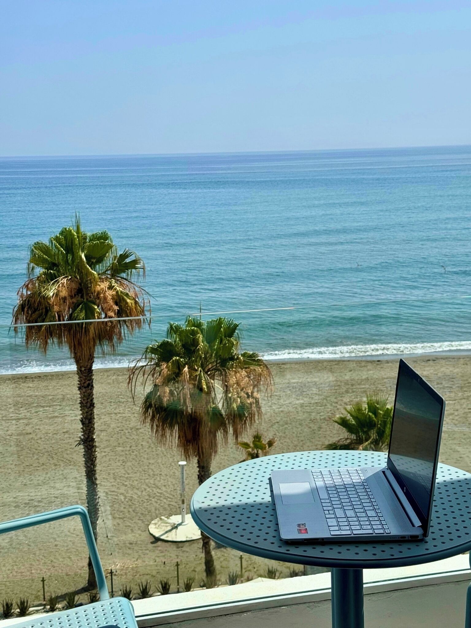 Un ordinateur portable est posé sur une table ronde, avec une vue sur la plage, des palmiers et un ciel dégagé.