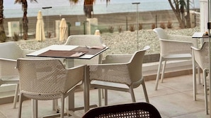An outdoor seating area with tables and chairs, overlooking a beach with palm trees and a clear sky.