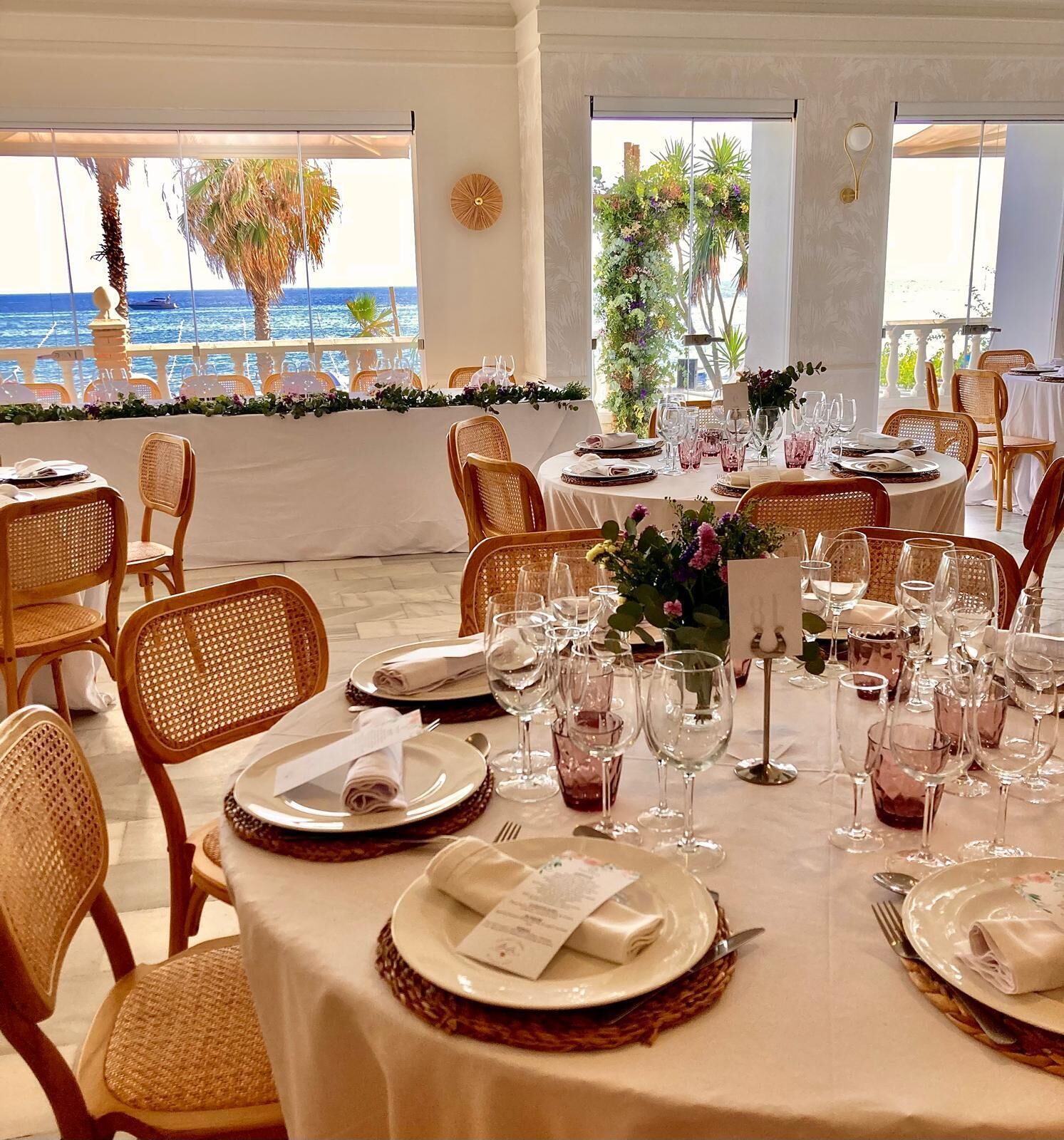 A dining area with round tables set for a meal, chairs, and a view of the sea.