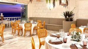 A dining area with wicker chairs, round tables, hanging plants, and a view of the sea.