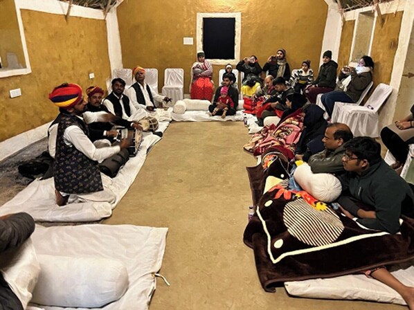A room with a long table set up for a meeting or event, with people seated on chairs and some on the floor. - Heartland Dunes (Jaisalmer)