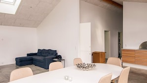 Ein modernes, minimalistisches Zimmer mit einer Eckcouch, einem Esstisch mit Stühlen, einem großen Fenster und einer Holzkassette.