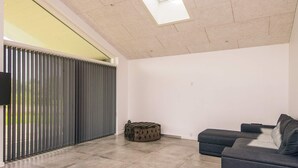 Ein modernes Wohnzimmer mit einem grauen Sofa, einem runden Hocker und einem großen Fenster mit Jalousien.