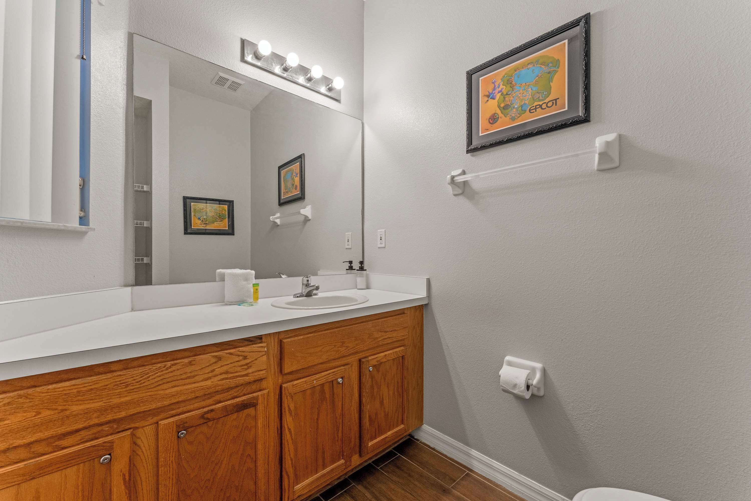 A bathroom with a wooden vanity, white sink, and a large mirror with lights. There are framed pictures on the wall and a map.