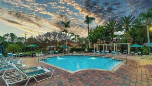 A swimming pool with lounge chairs and umbrellas, surrounded by palm trees and buildings. - Magic Getaway at Emerald Island (Kissimmee)