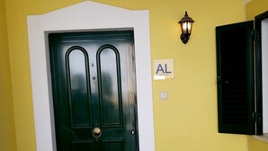 Eine schwarze Tür mit Messingbeschlägen, eine Wandleuchte und ein Schild mit den Buchstaben "AL" an einer gelben Wand.