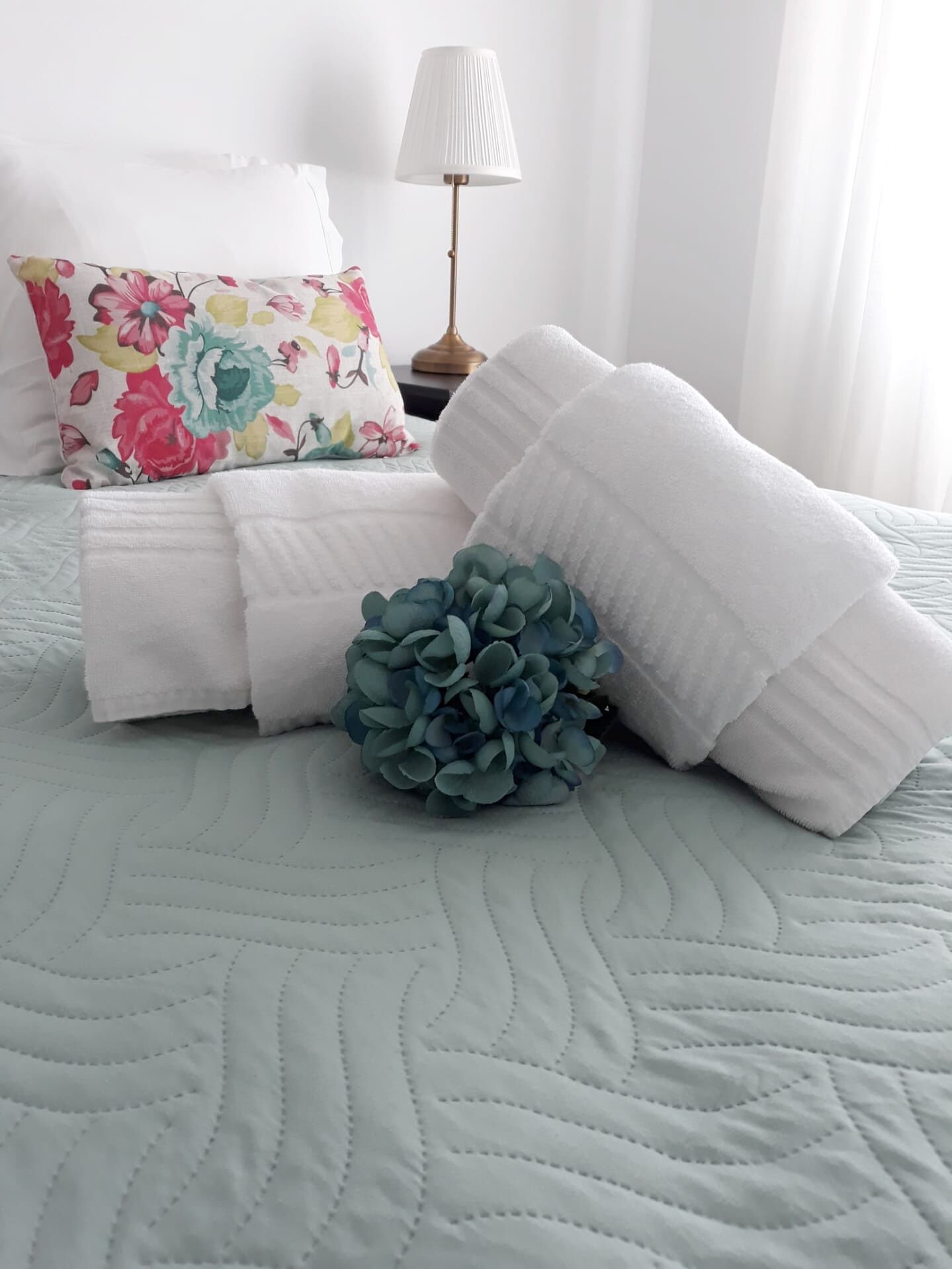Cama arrumada com toalhas brancas, almofada floral, planta azul decorativa e abajur.