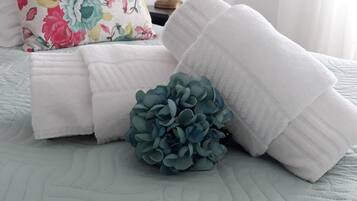 Cama arrumada com toalhas brancas, almofada floral, planta azul decorativa e abajur.