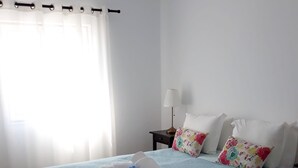 Ein ordentlich bezogenes Bett mit einer blauen Bettdecke, zwei weißen Kissen und einem Kissen mit Blumenmuster. Auf dem Nachttisch neben dem Bett stehen eine Lampe und ein gefaltetes Handtuch.