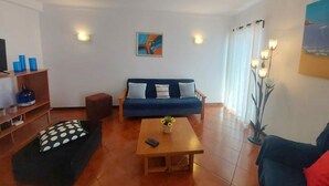 Ein Wohnzimmer mit einem blauen Sofa, einem Holztisch, einem schwarzen Hocker mit gepunktetem Kissen. Auf einem kleinen Tisch stehen eine Blumenvase und eine Fernbedienung.