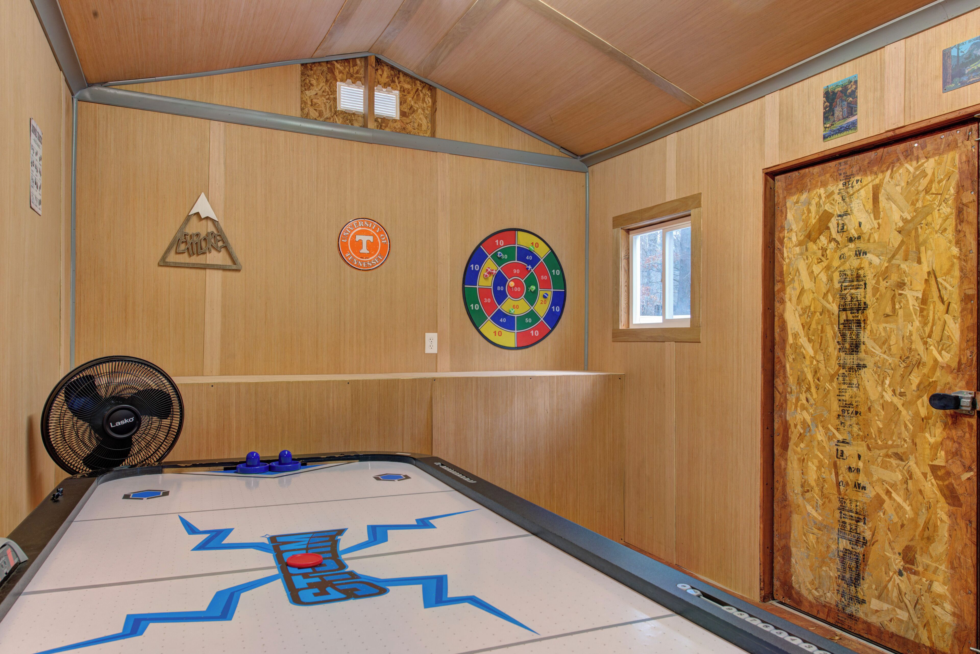 Una habitación con mesa de juego tipo bolera, paredes de madera, un ventilador y una puerta con ventana.