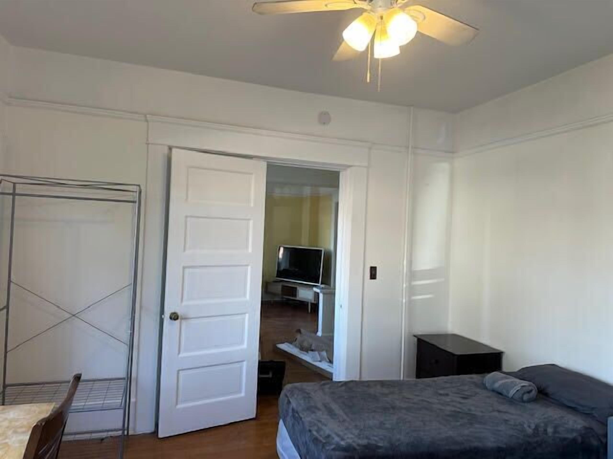 A bedroom with a bed, a ceiling fan, a TV, and a door leading to another room.