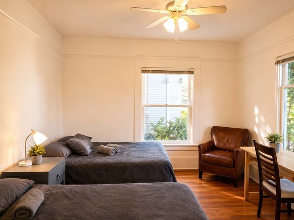A bedroom with two beds, a ceiling fan, a window with blinds, a desk with a chair, and a potted plant.