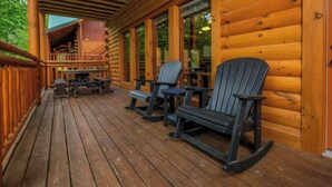 Terraza de madera con sillas Adirondack y una mesa, con vista al interior de una cabaña.