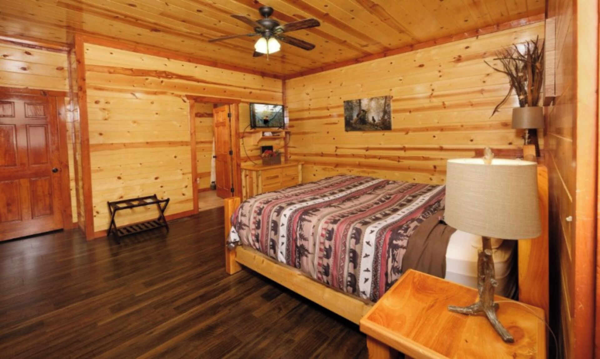 Une chambre dans un chalet en bois, avec un lit, un ventilateur de plafond, un téléviseur et un tableau accroché au mur.