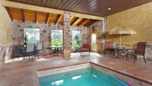 Una piscina interior con paredes de piedra y vigas de madera, rodeada de asientos exteriores y una puerta que da al exterior.