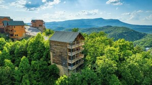 A wooden cabin with a balcony, surrounded by dense forest and mountains in the background.