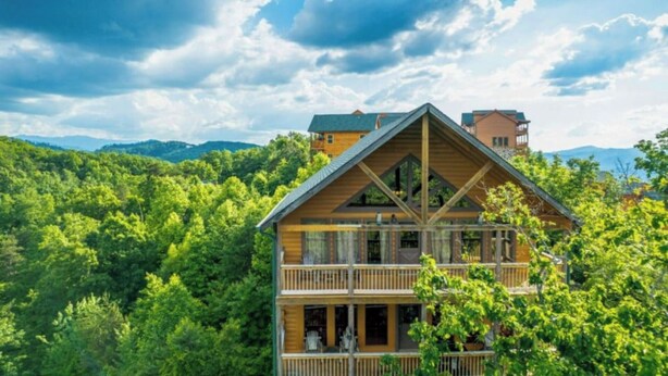 A two-story wooden cabin with a balcony, surrounded by lush greenery and mountains in the background.