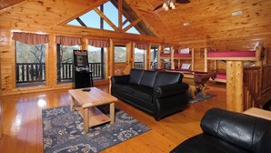 A wooden cabin interior with a wooden floor, a black leather sofa, a wooden coffee table, a pool table, and a view of the outdoors through large windows.