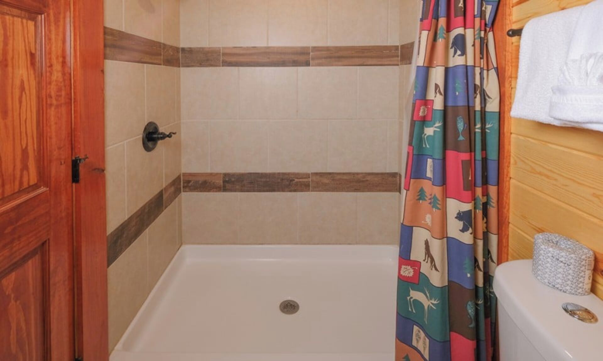 A shower area with a wooden door, beige tiles, and a colorful shower curtain.