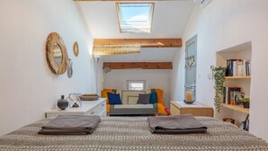 Un dormitorio con tragaluz, una cama con muchos almohadones, una mesita de noche con lámpara, un espejo y una estantería.
