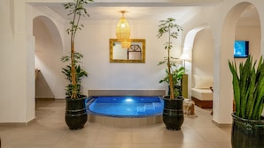 A modern indoor swimming pool with a blue tiled floor, surrounded by white walls and arches. There are two large potted plants on either side of the pool, and a large mirror above it. - PRIVATE RIAD DAR TOURIA (Marrakesh)