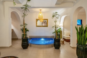 A modern indoor swimming pool with a blue tiled floor, surrounded by white walls and arches. There are two large potted plants on either side of the pool, and a large mirror above it. - PRIVATE RIAD DAR TOURIA (Marrakesh)