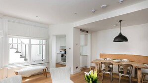 Un vaste espace de vie comprenant une table à manger, des fauteuils blancs et un banc en bois. Un luminaire suspendu noir est accroché au-dessus de la table.