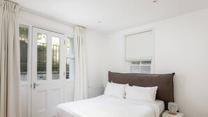 Une chambre à coucher avec un grand lit, des rideaux blancs, une porte blanche et une fenêtre donnant sur un paysage verdoyant.