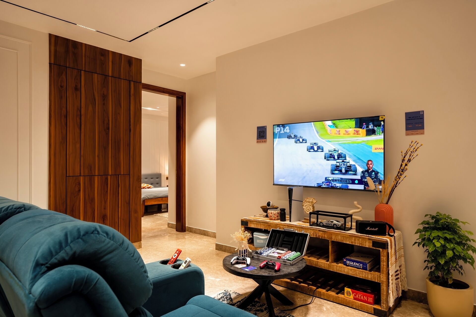 Un soggiorno con una grande TV a schermo piatto che mostra un gioco di corse, un divano blu, un tavolino da caffè con joystick e un mobiletto in legno con oggetti decorativi.