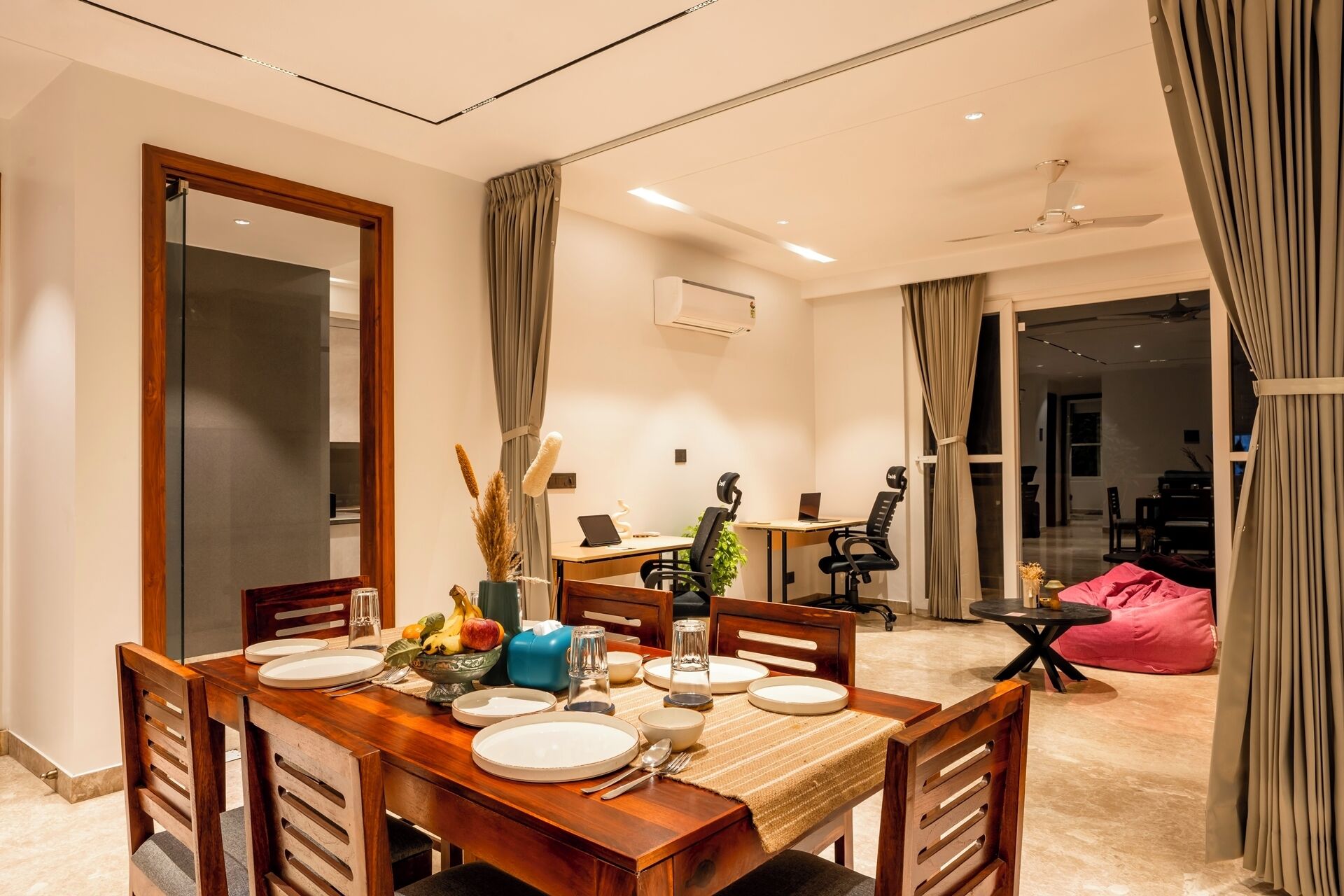 Un comedor con una mesa de madera dispuesta para cuatro, un espejo y vistas a una sala de estar con sofá y televisor.