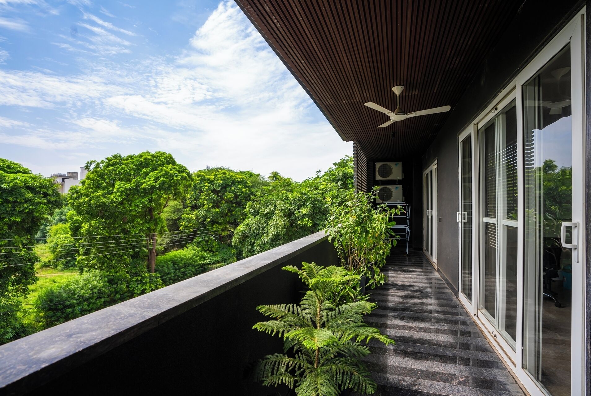 A balcony with a view of lush greenery and a modern building with large windows and a ceiling fan.