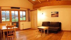 A cozy living room with a wooden floor, a sofa, a table, and a window with a view of a house and greenery. - Matthiasgut - Tauernview S (Radstadt)